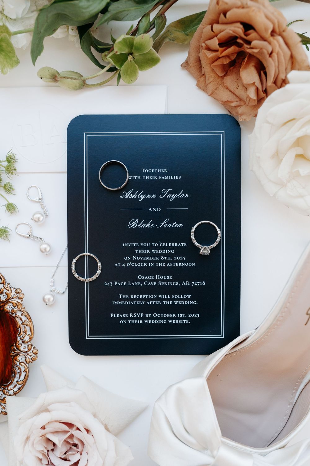 Elegant navy blue wedding invitation for Ashlynn Taylor and Blake Soter surrounded by pearl jewelry, rings, and blush roses.