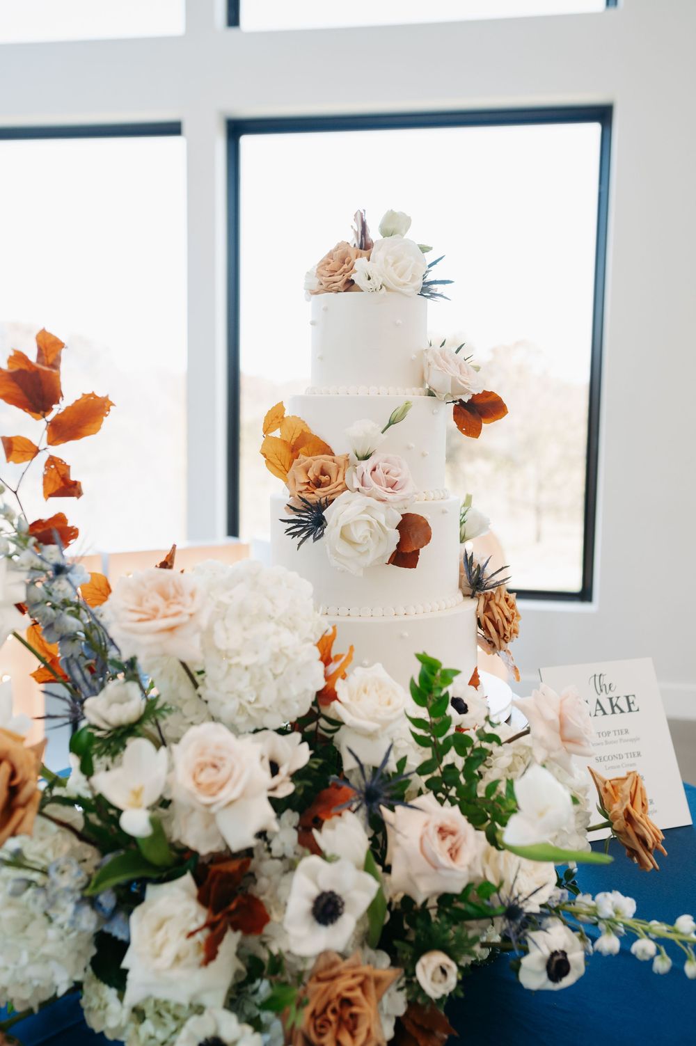 Elegant four-tier white wedding cake adorned with blush, orange, and white florals, surrounded by lush floral arrangements on a blue table.