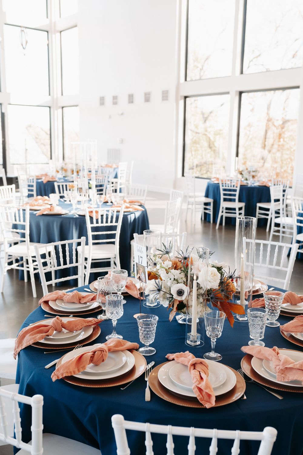 Elegant wedding reception table with navy blue linen, blush pink napkins, gold chargers, crystal glasses, and floral centerpiece.