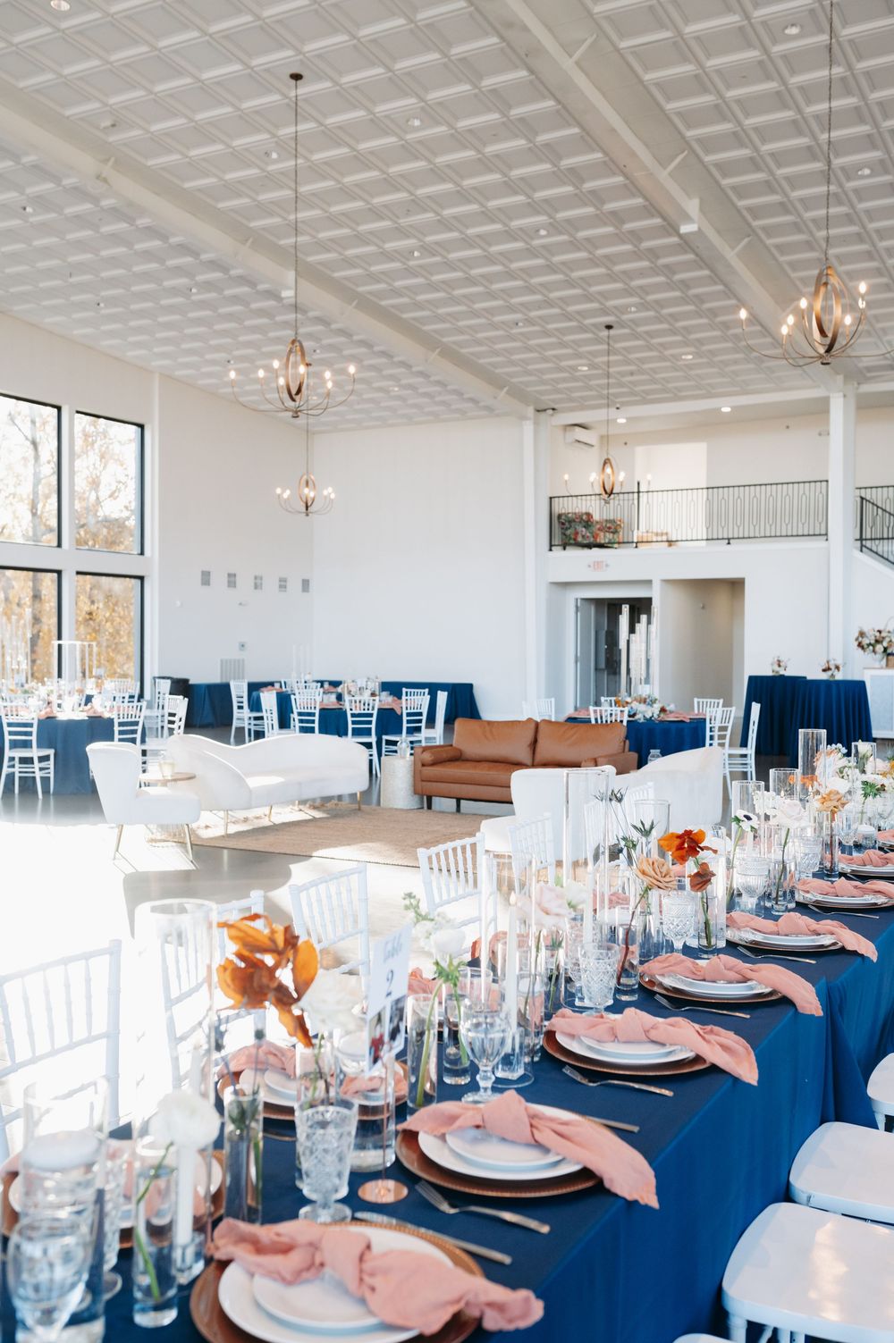 Elegant wedding reception with navy blue tablecloths, blush pink napkins, white chiavari chairs, and gold pendant lights in a bright venue.