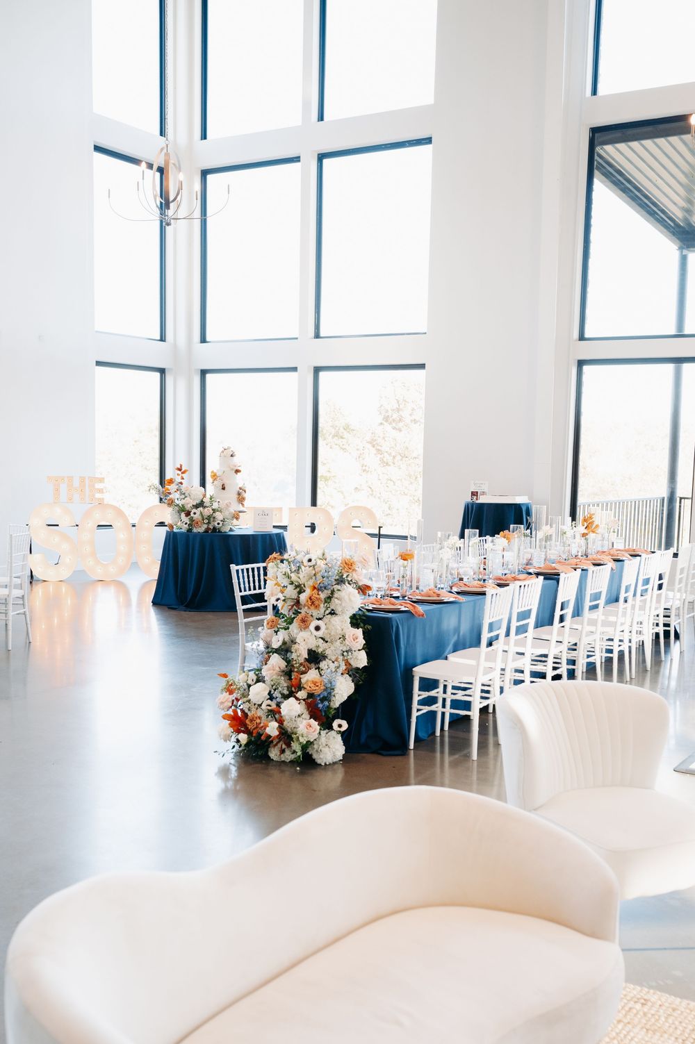 Elegant wedding reception hall with navy blue tablecloths, white chairs, floral arrangements, and floor-to-ceiling windows.