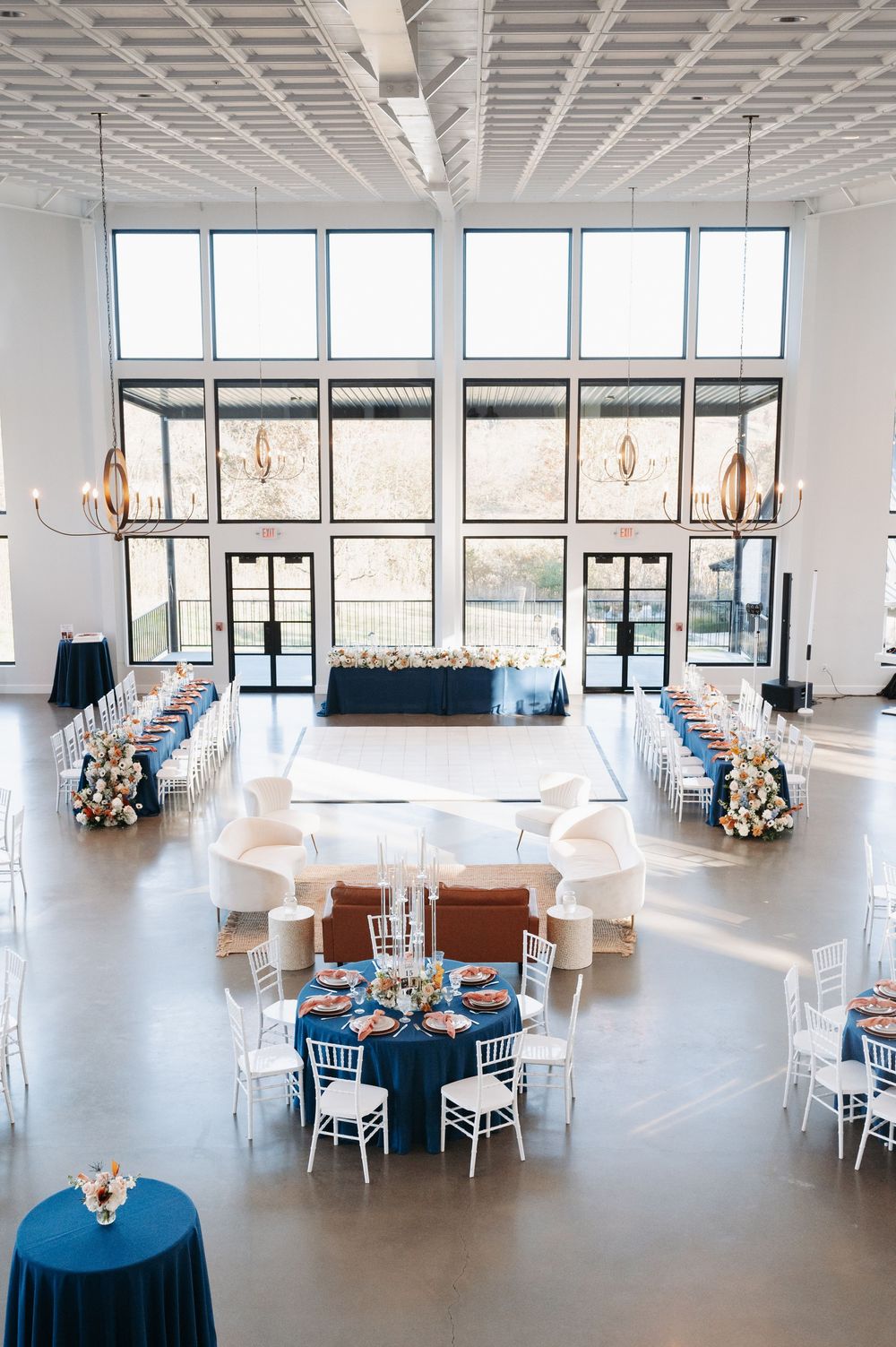Spacious modern wedding venue with navy blue round tables, white chiavari chairs, and large floor-to-ceiling windows.