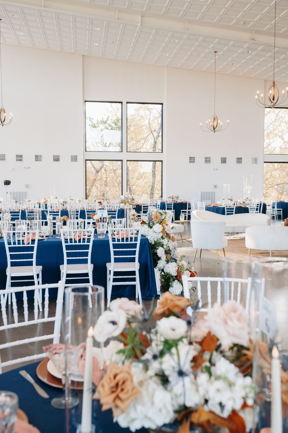Elegant wedding reception hall with navy blue tablecloths, white chiavari chairs, floral centerpieces, and large windows with natural light.