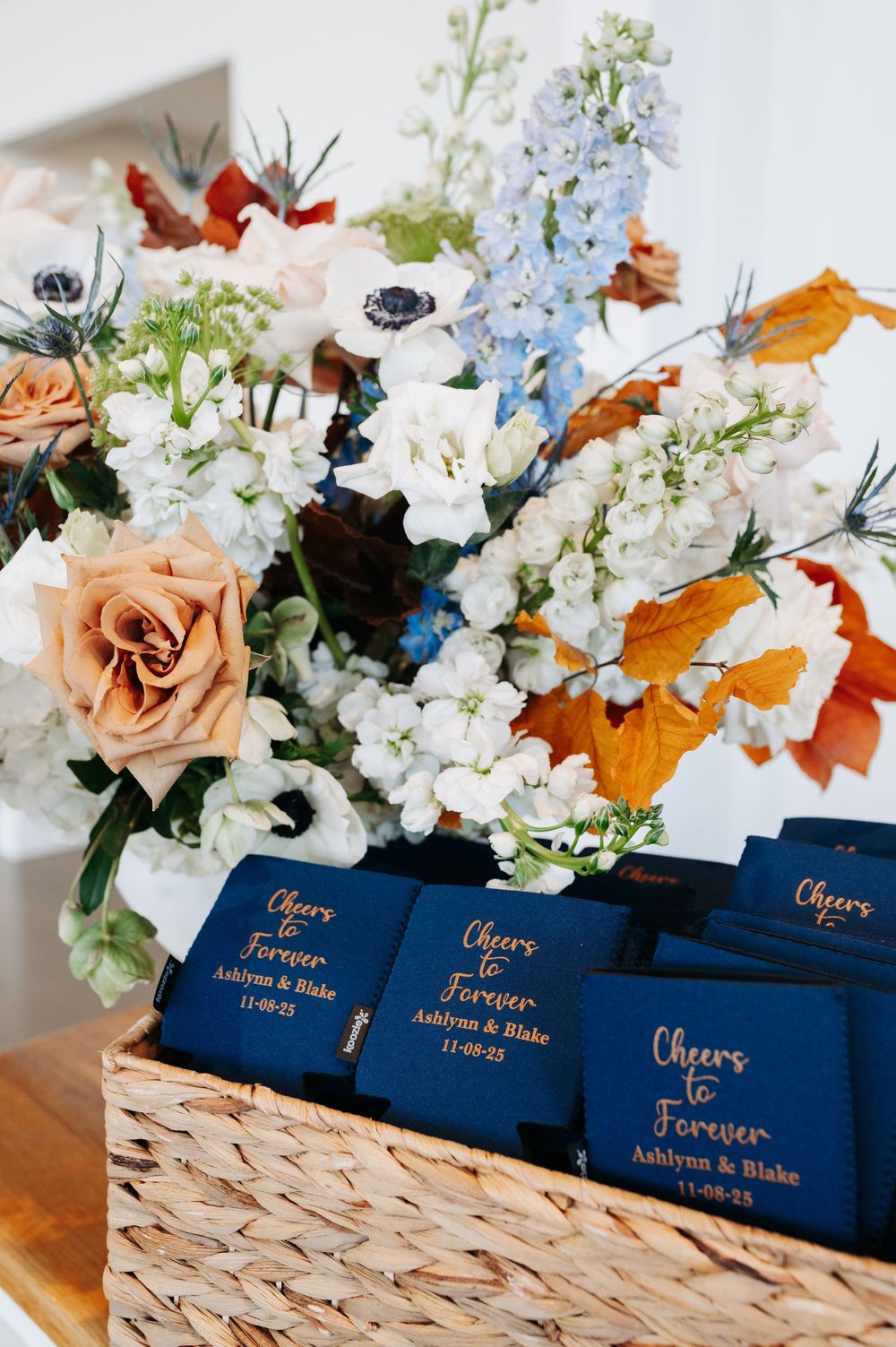 Navy blue wedding koozies reading Cheers Forever Ashlynn and Blake beside a floral arrangement of roses and autumn leaves.