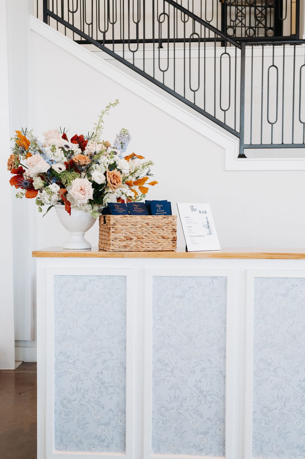 White reception desk with blue floral panels, topped with a colorful flower arrangement, wicker basket, and signage near a staircase.