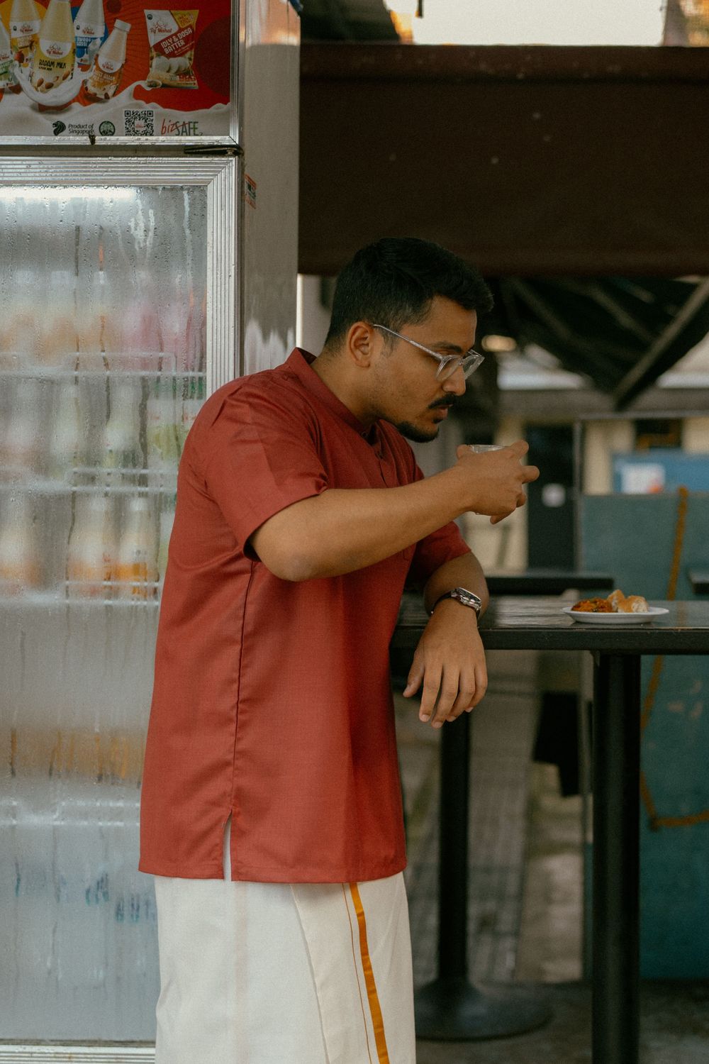 Man in red shirt and white dhoti eating at an outdoor food stall, wearing glasses and a watch.