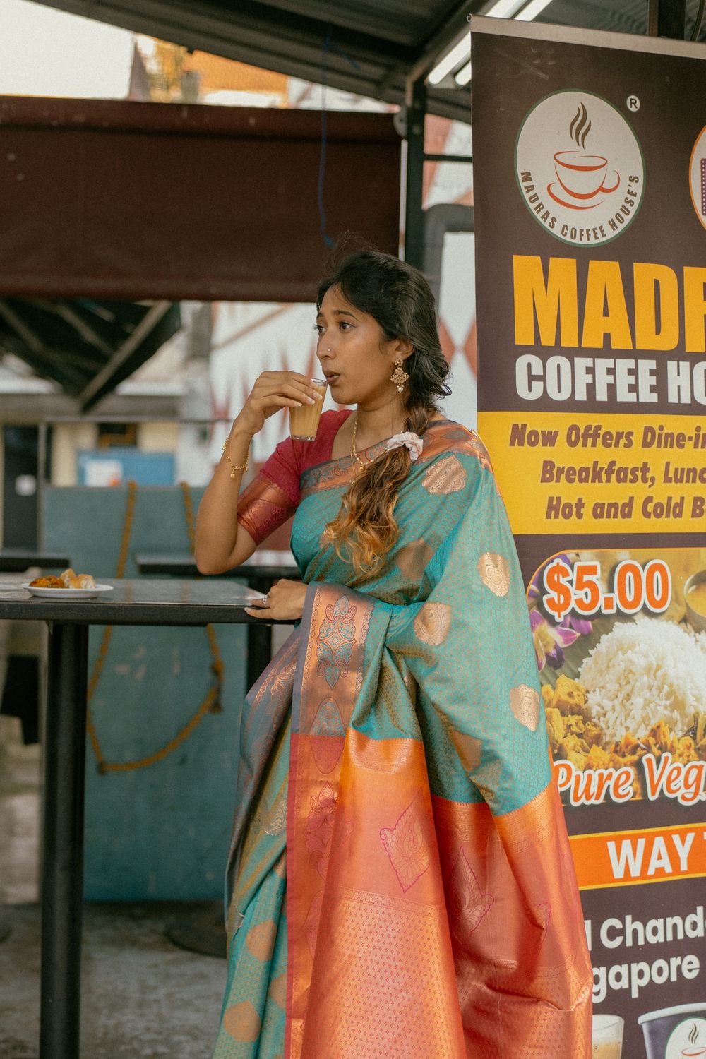 Woman in teal and orange silk saree sipping a drink at Madras Coffee House in Singapore.
