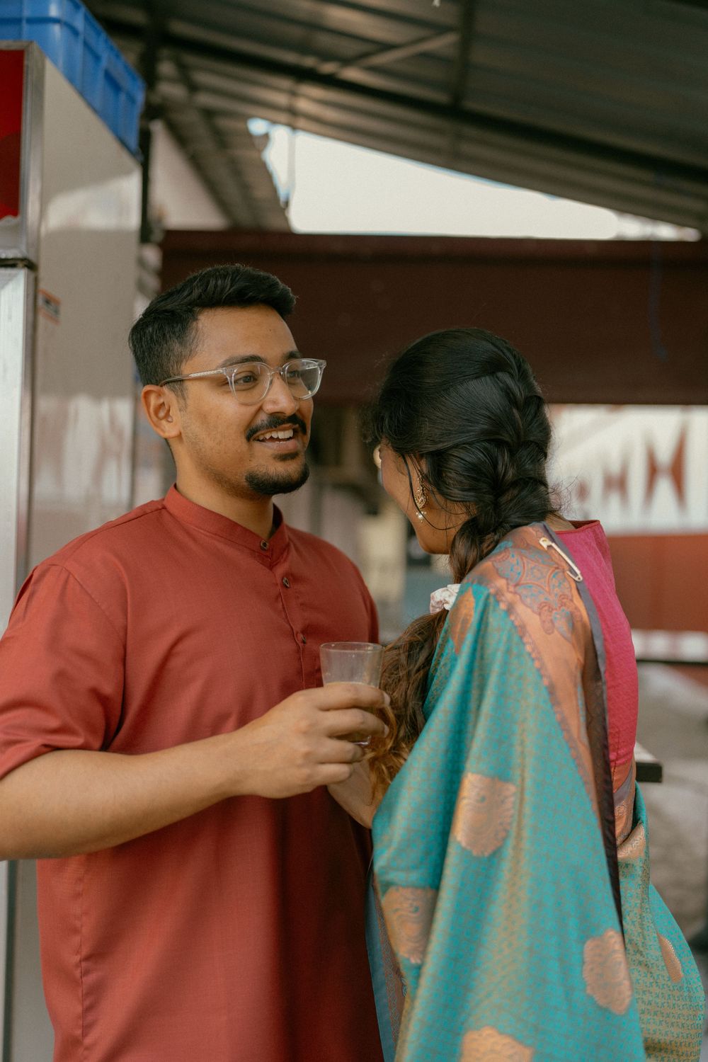 Indian couple in traditional attire conversing outdoors, man in red kurta holding drink, woman in teal and red saree.