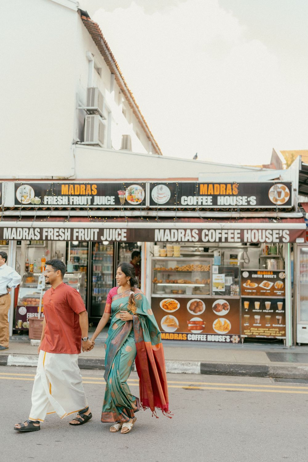 Indian couple in traditional attire walking past Madras Fresh Fruit Juice and Coffee House storefront.