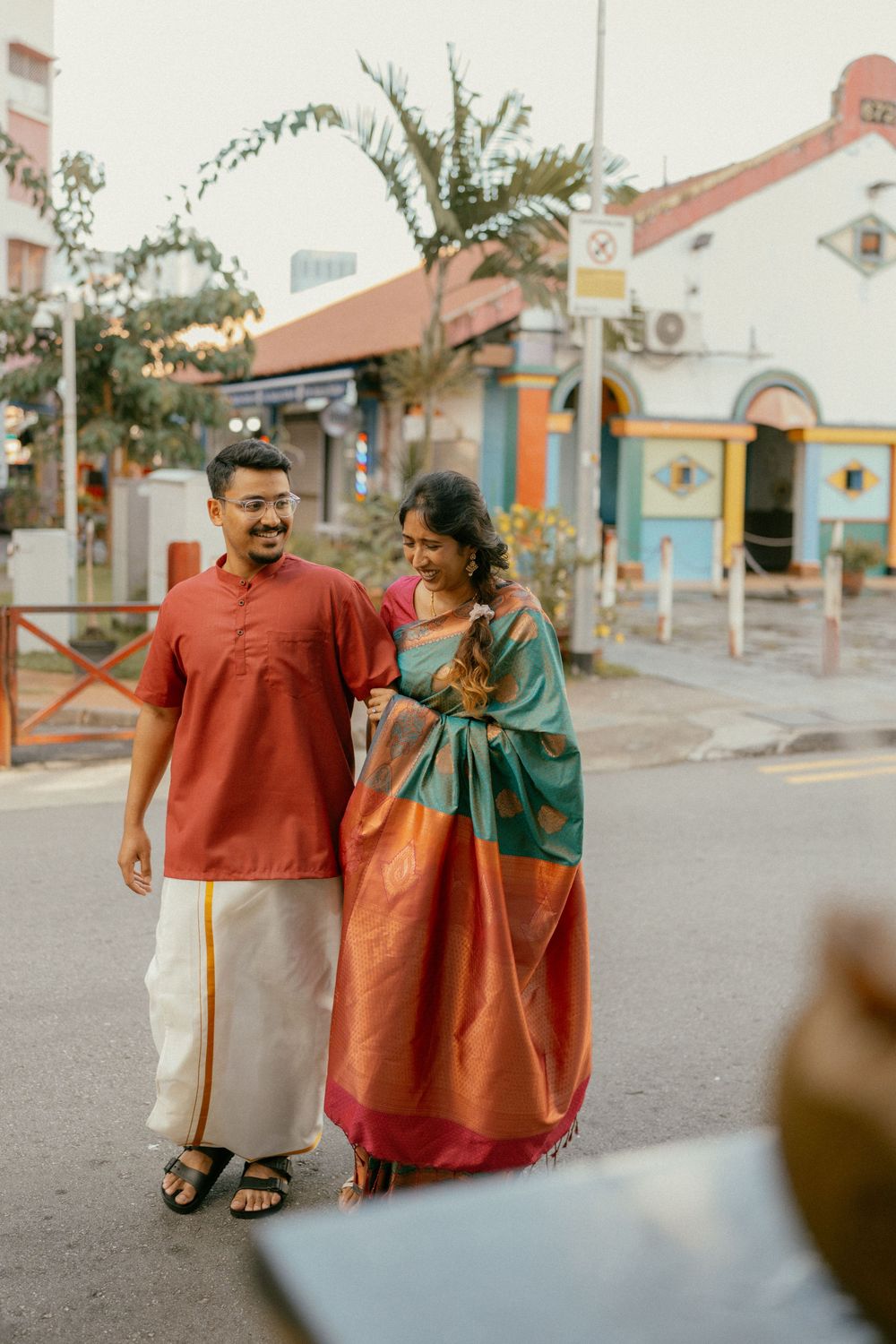 Indian couple in traditional attire walking on a street, man in red kurta and dhoti, woman in green and red saree.