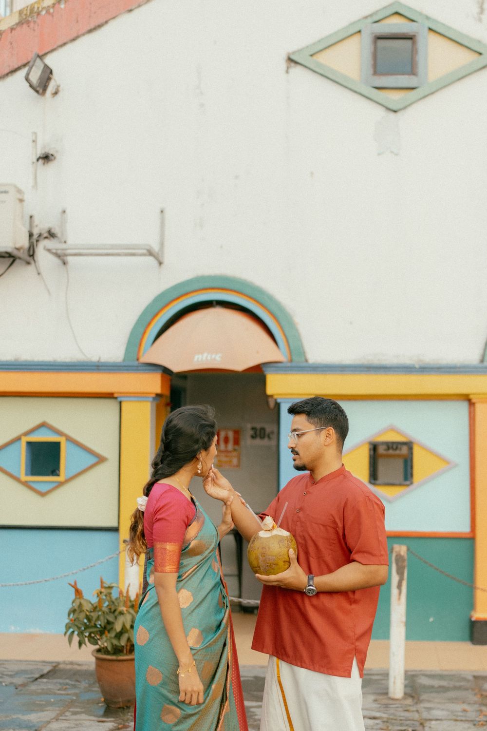 Indian couple in traditional attire sharing a coconut drink outside a colorful temple building.