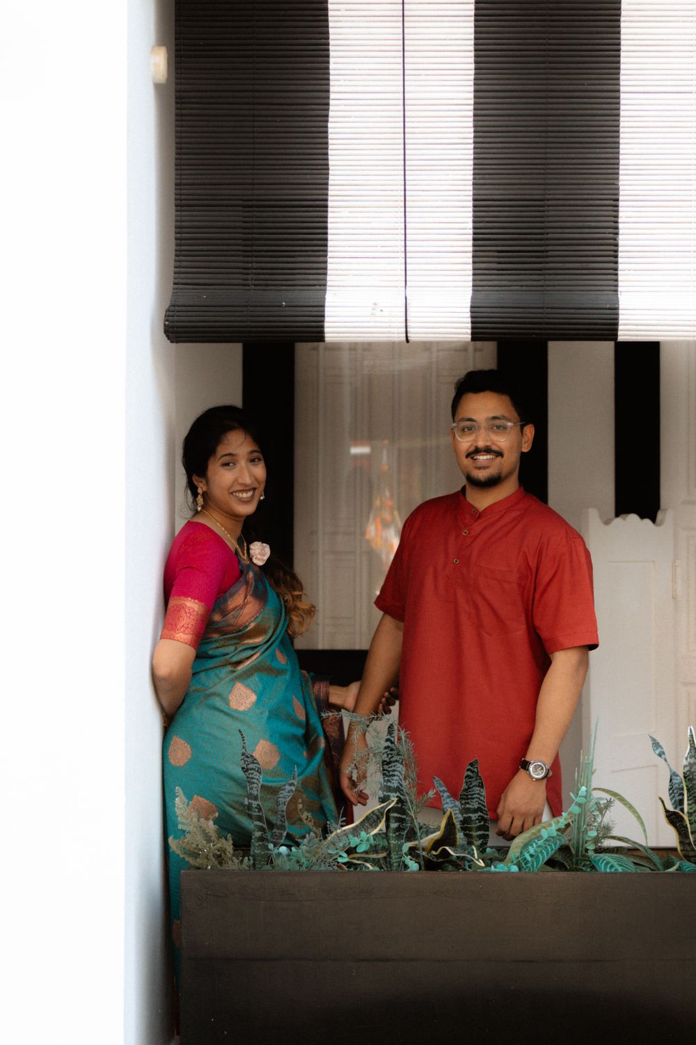 A smiling couple in traditional Indian attire standing by a balcony planter, woman in teal saree, man in red kurta.