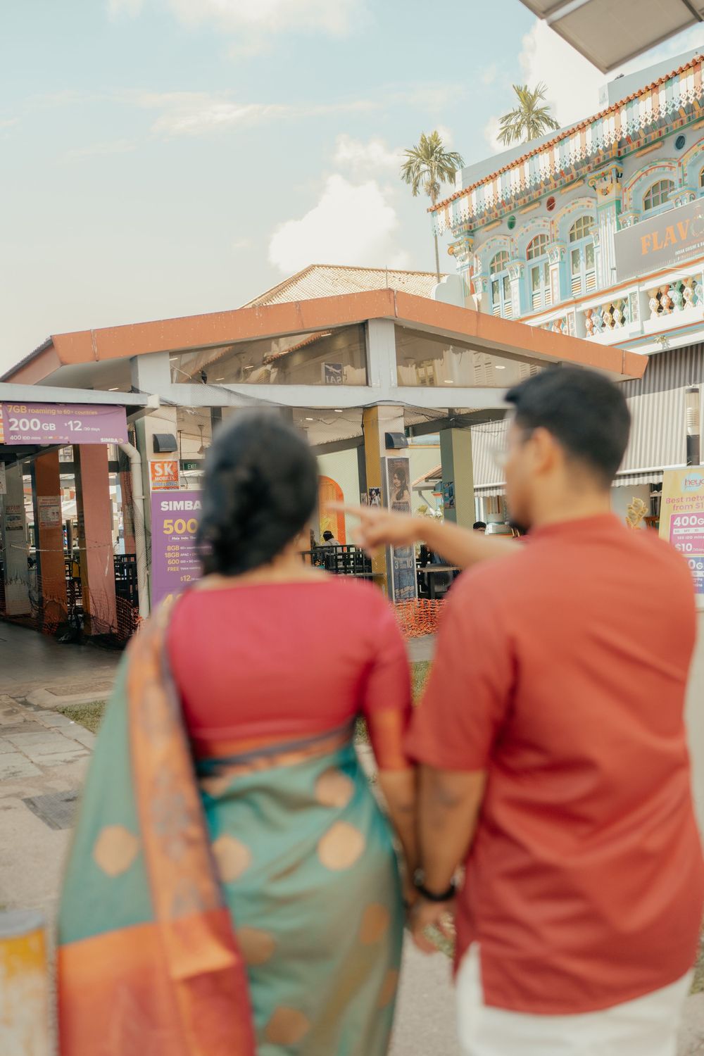 Indian couple in traditional attire holding hands, viewed from behind, exploring a colorful heritage building outdoors.