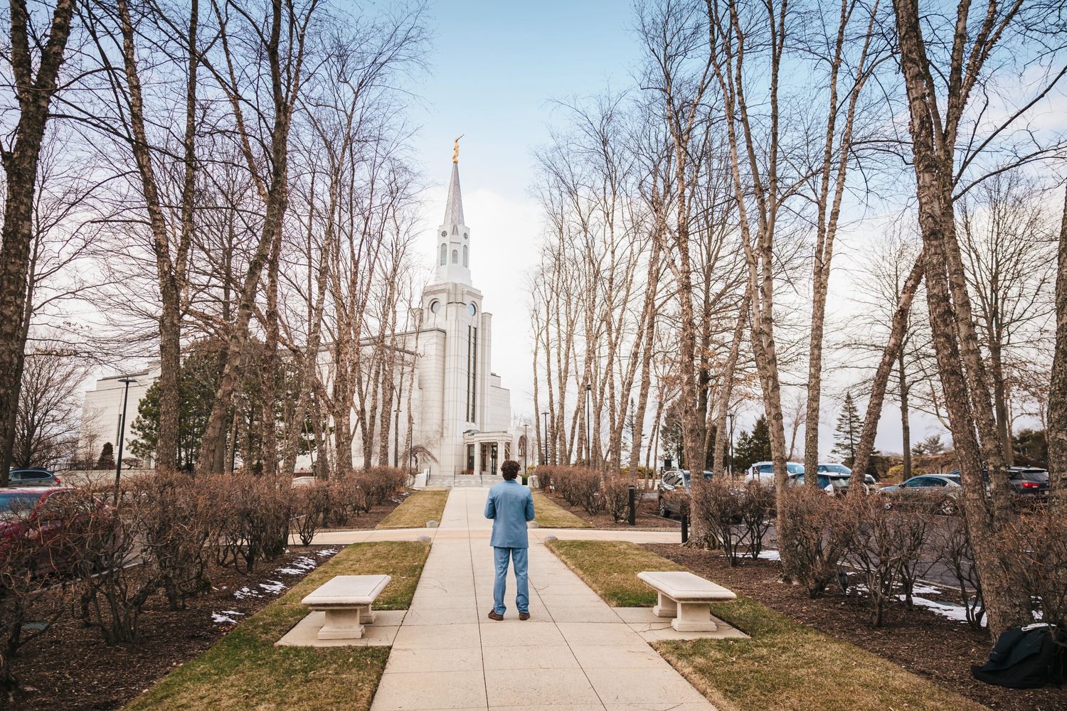 The groom standing alone on a tree-lined pathway facing Boston Temple  in the distance.  Elopement at Boston Massachusetts Temple, Belmont,