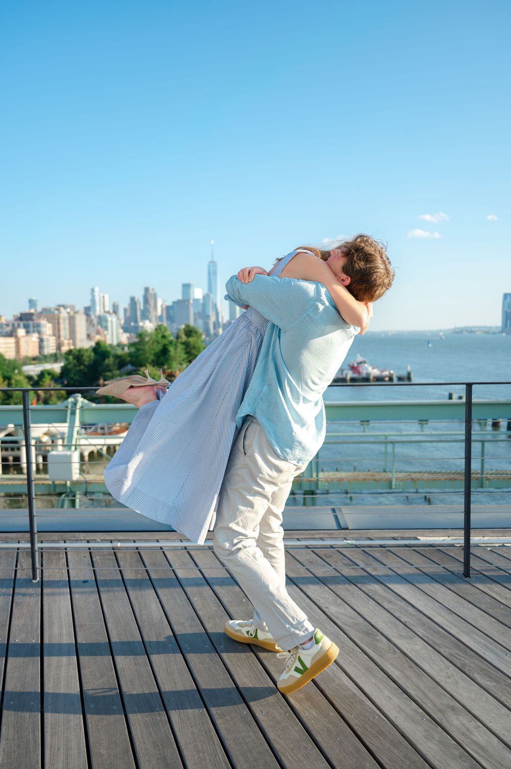Couple sharing a romantic lift embrace on a rooftop deck with NYC skyline in background.