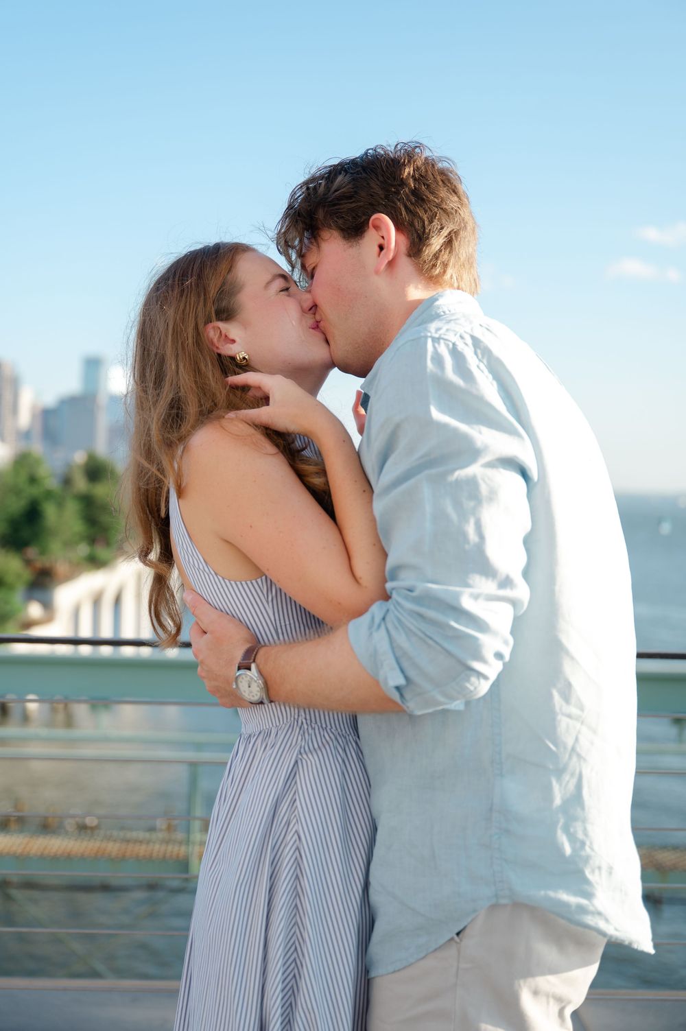 A couple shares a romantic kiss outdoors with a city skyline in the background on a sunny day.