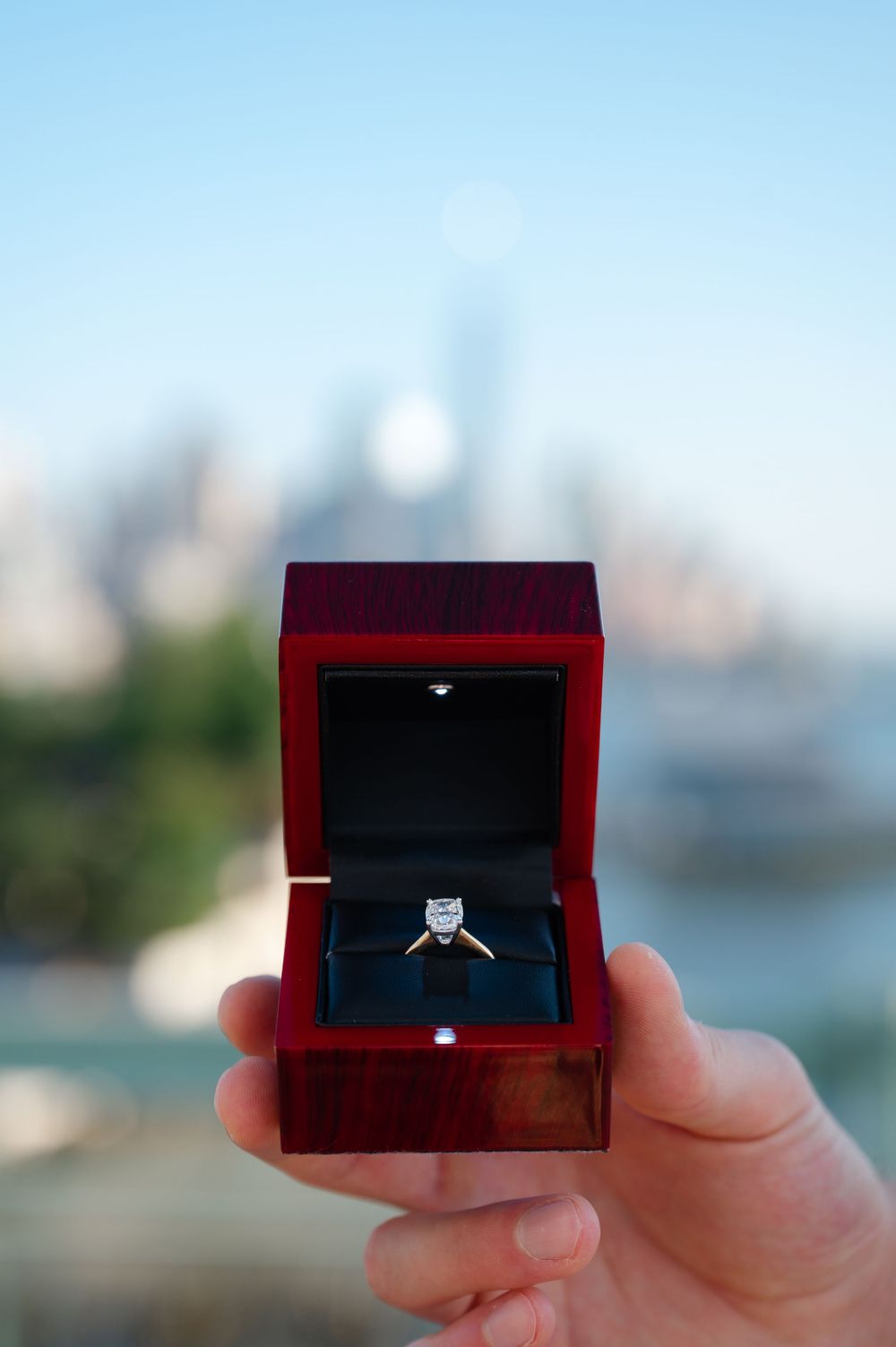 Diamond solitaire engagement ring in red wooden box with NYC skyline background.