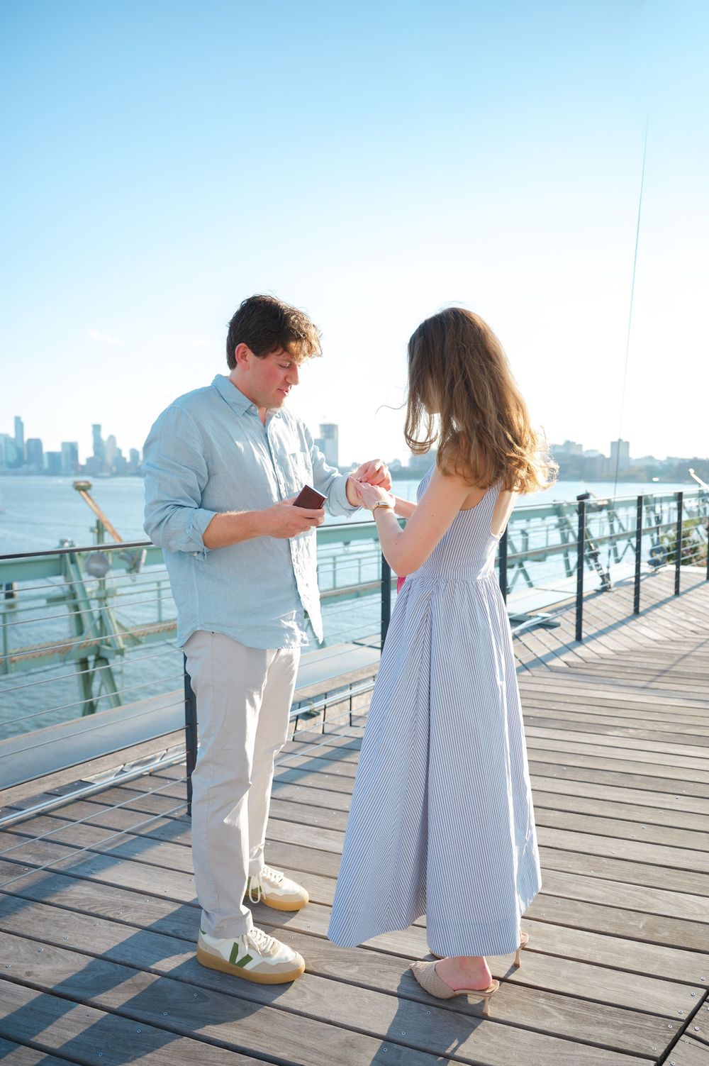 Man placing engagement ring on womans finger on a sunny waterfront pier with city skyline backdrop.