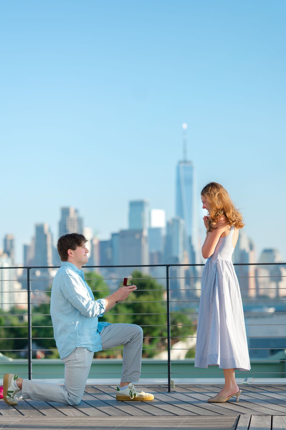 Man proposes on one knee with ring box on rooftop terrace with NYC skyline and One World Trade Center backdrop.
