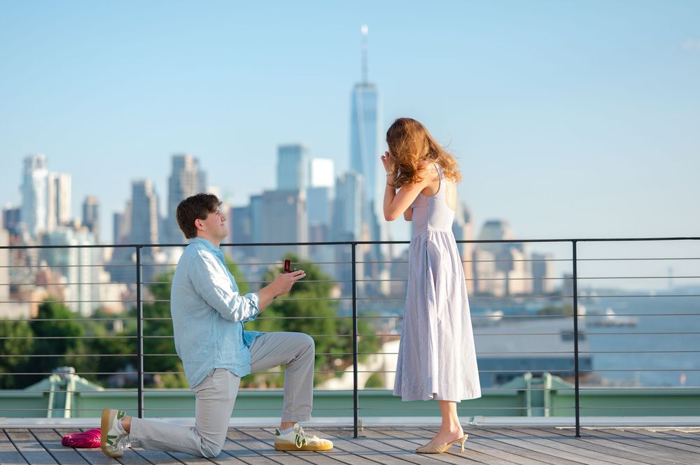 Man proposes on rooftop with NYC skyline backdrop, woman reacts with surprise and joy during romantic engagement moment.