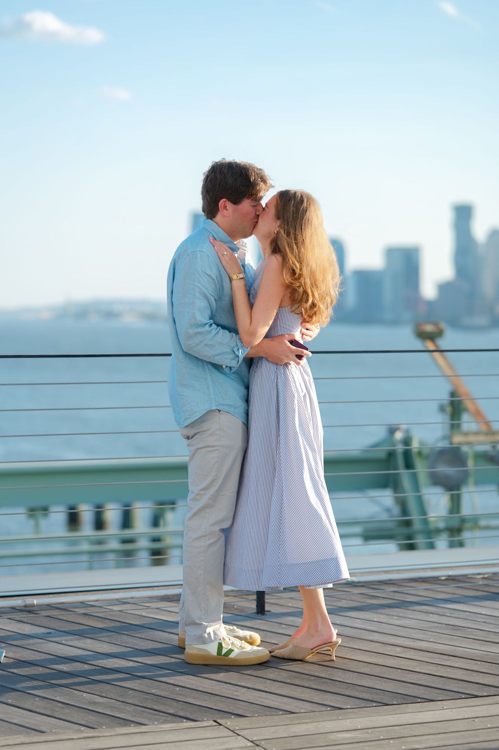 Couple sharing romantic kiss on waterfront deck with city skyline behind them.