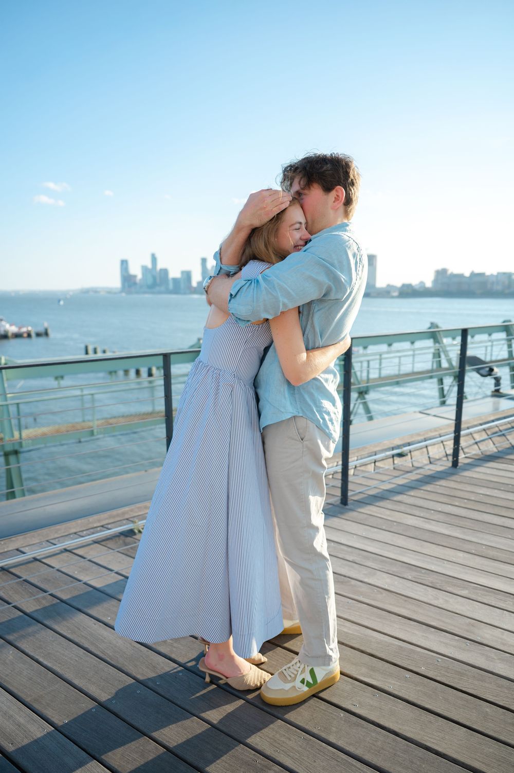 Couple sharing a tender embrace on a waterfront deck with city skyline and blue sky in background.