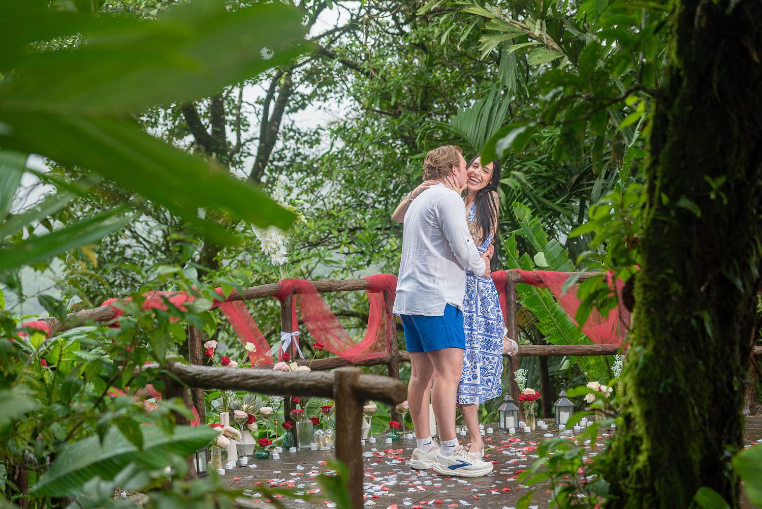 Couple sharing a romantic kiss on a red bridge surrounded by lush tropical greenery in a garden setting.