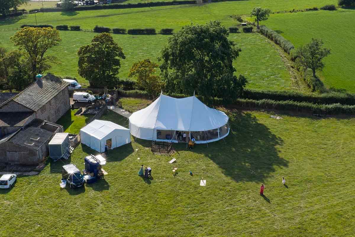 Aerial view of a large white oval marquee tent set up in a green field next to a stone building for an outdoor event.