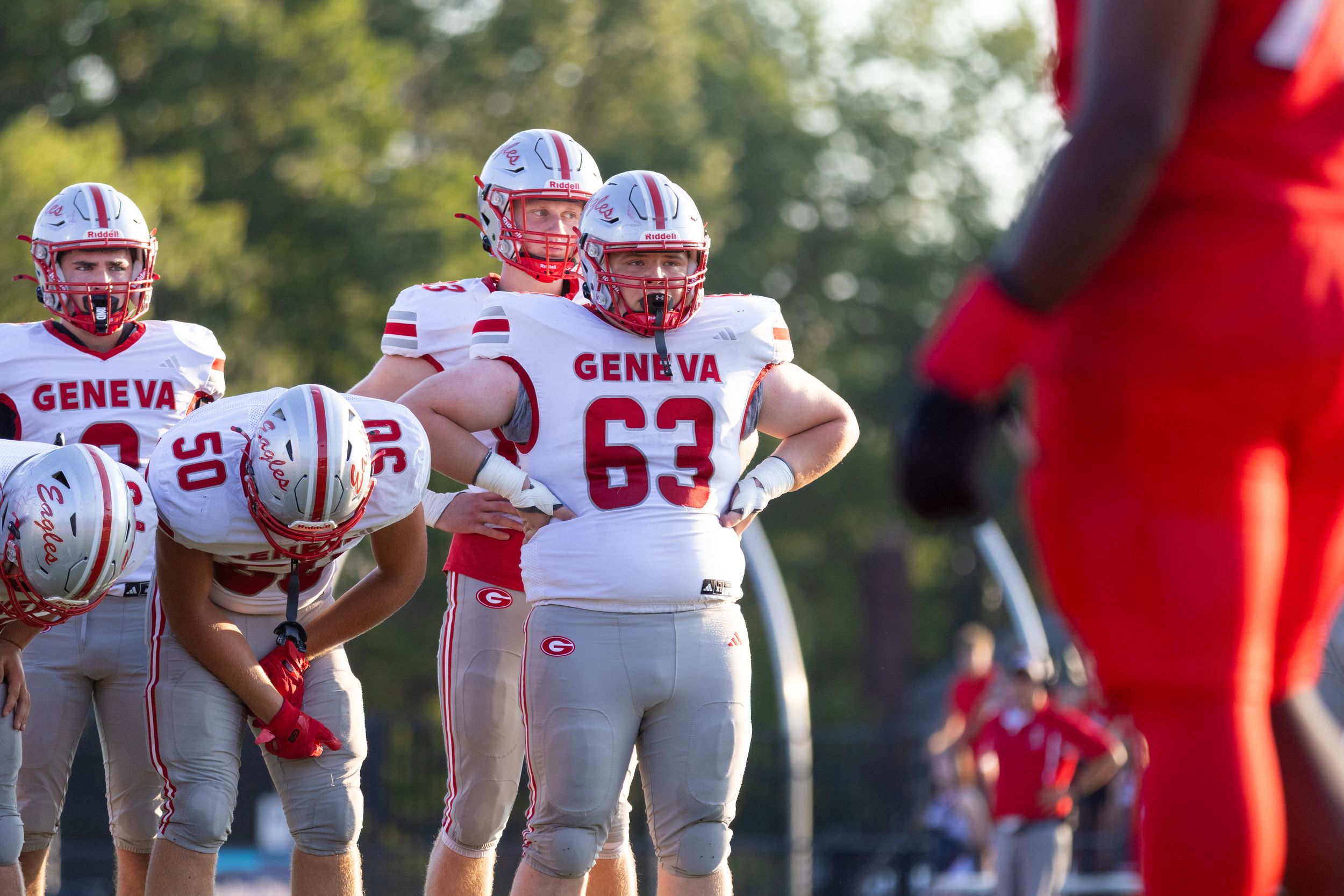 Geneva football players in white and red uniforms stand on the field during a game, with player 63 in focus.