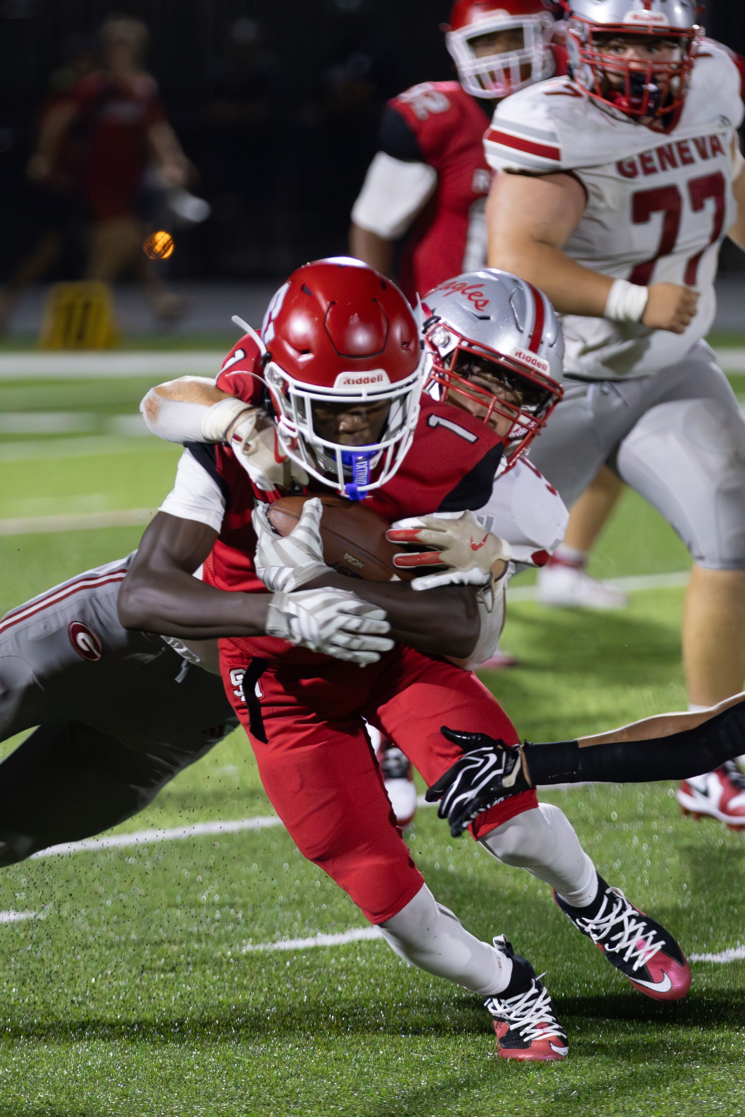 High school football players in red uniforms tackle a ball carrier during a night game on a green field.