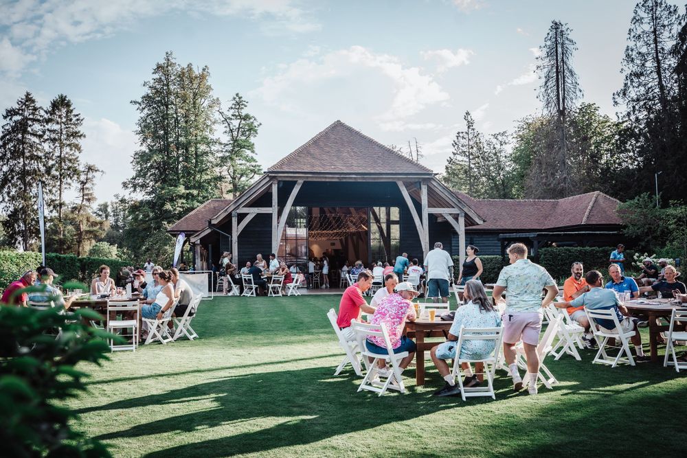Guests enjoy an outdoor summer event on lush green lawn with white chairs near a rustic open-air pavilion.