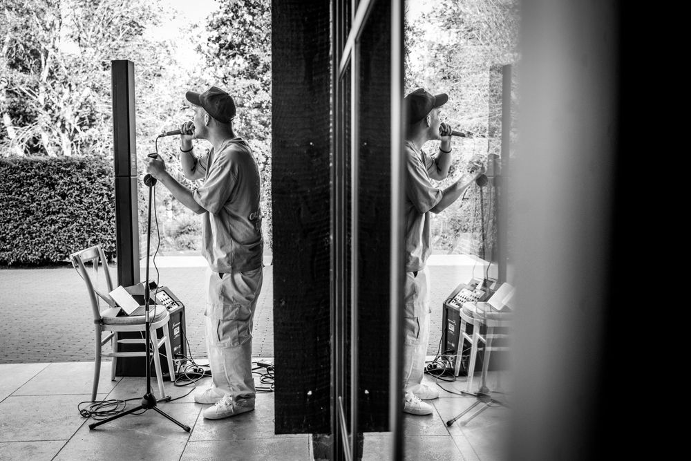 Black and white photo of a singer performing with a microphone, reflected in glass doors at an outdoor venue.