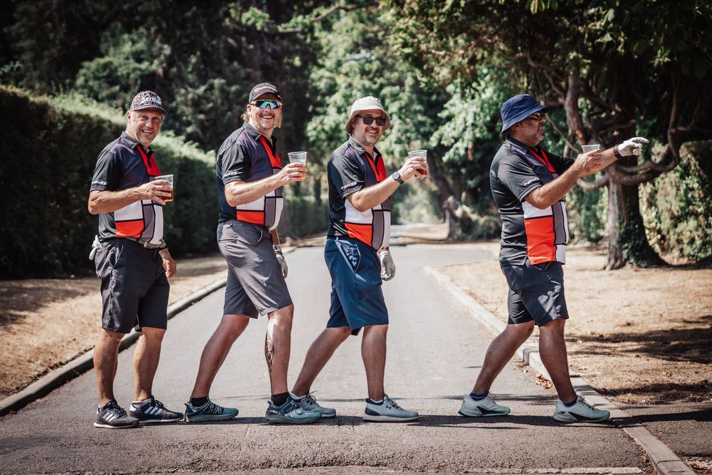 Four men in matching cycling jerseys and shorts walk in a line on a sunny tree-lined path, each holding drinks.