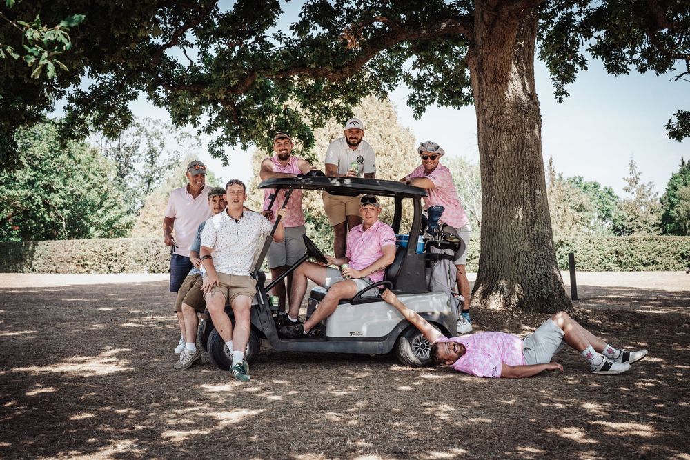 Group of men posing with a golf cart under a large tree on a sunny golf course.