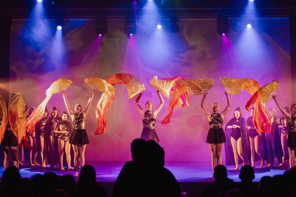 Dance performers in black costumes hold large decorative fans under colorful stage lighting during a show.