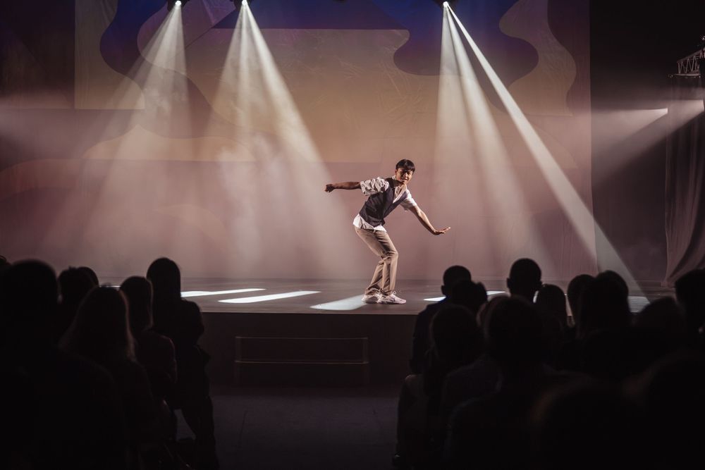 Solo performer dances on a spotlit stage before a silhouetted audience in a darkened theater.