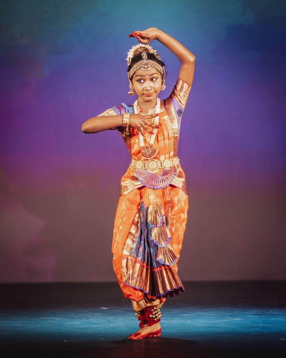 Young girl performs classical Bharatanatyam dance in vibrant traditional orange costume on a lit stage.