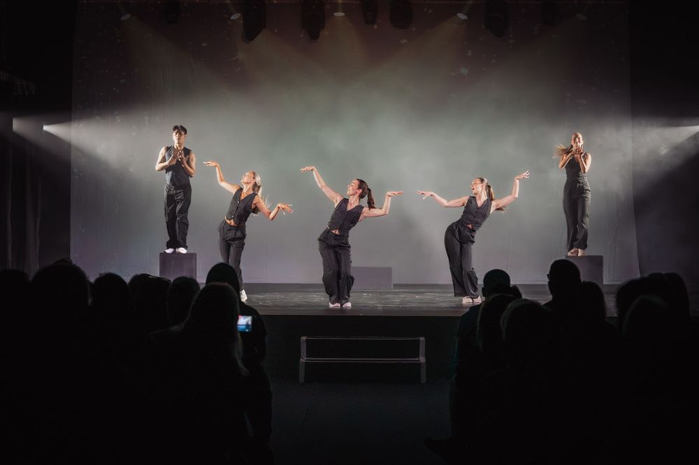 Five dancers in black perform synchronized choreography on a dramatic stage with atmospheric lighting and fog.