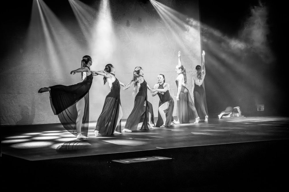 Black and white photo of elegant dancers performing on a dramatic stage with spotlights and smoke effects.