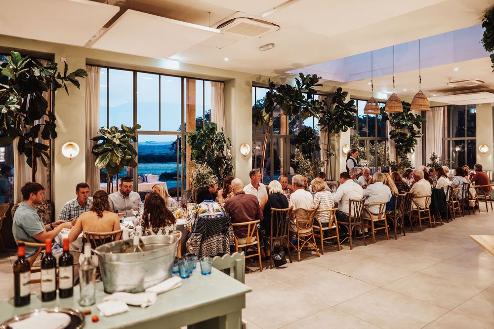 Guests dine together at long tables in a bright modern restaurant with large windows and lush plants.