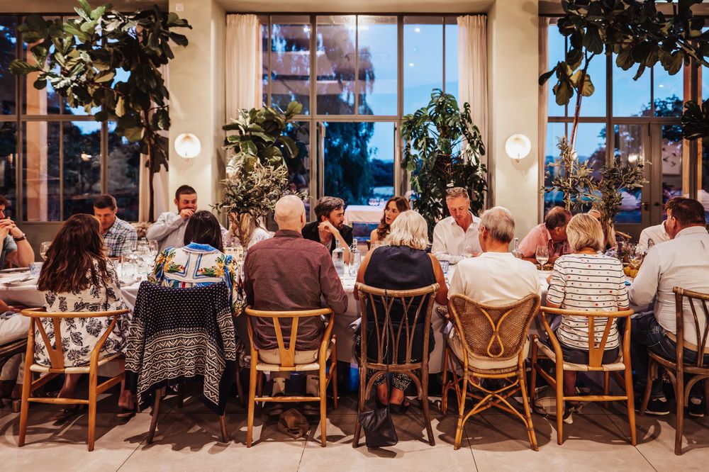 Group of guests seated around a large dining table in an elegant restaurant with lush indoor plants.