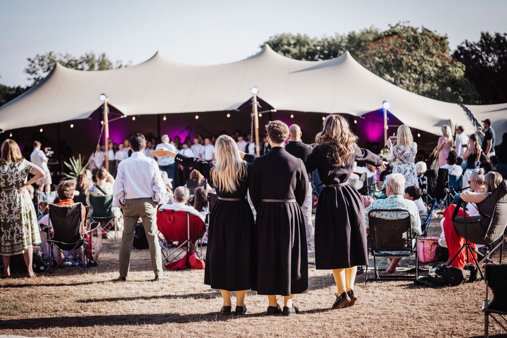 Outdoor festival event with guests gathered near a large white marquee tent with string lights and purple stage lighting.
