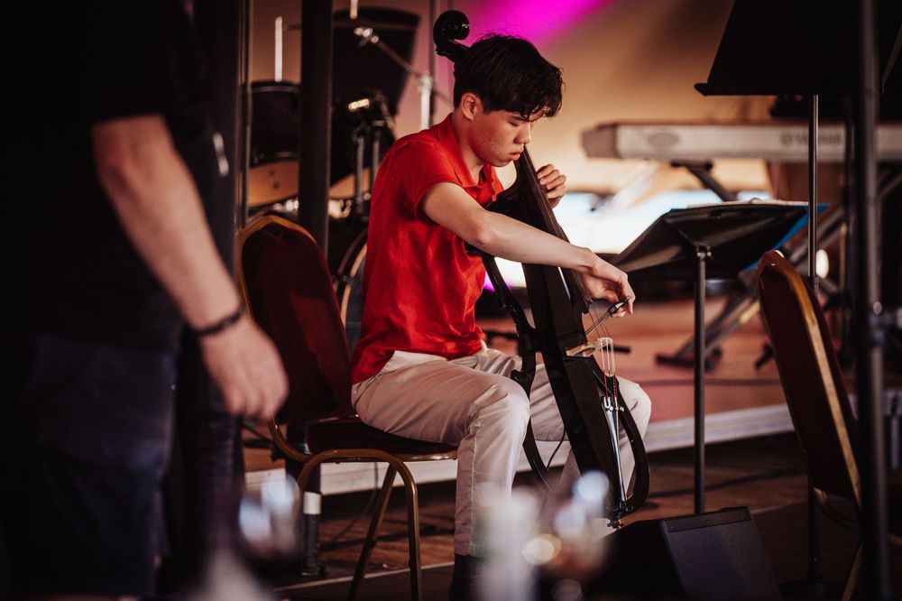 Young male musician in red shirt plays cello on stage during a live performance under dramatic lighting.