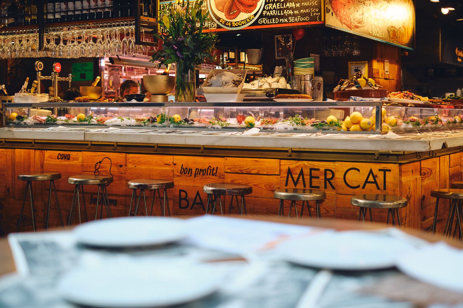 Warm interior of Mercat Bar with wooden counter, bar stools, and food display in a bustling market setting.