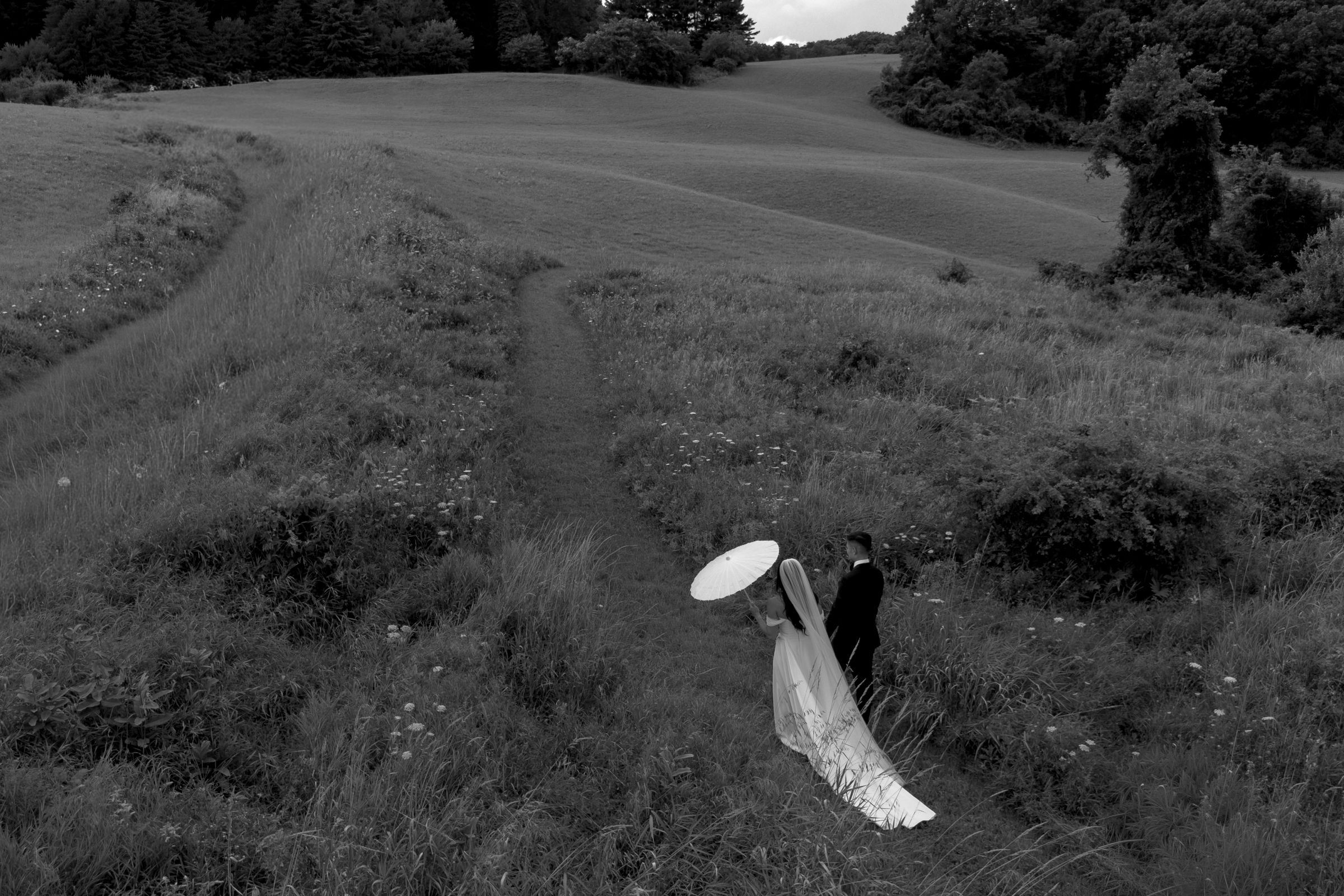 Black and white aerial photo of a couple in wedding attire walking through a lush green countryside field.