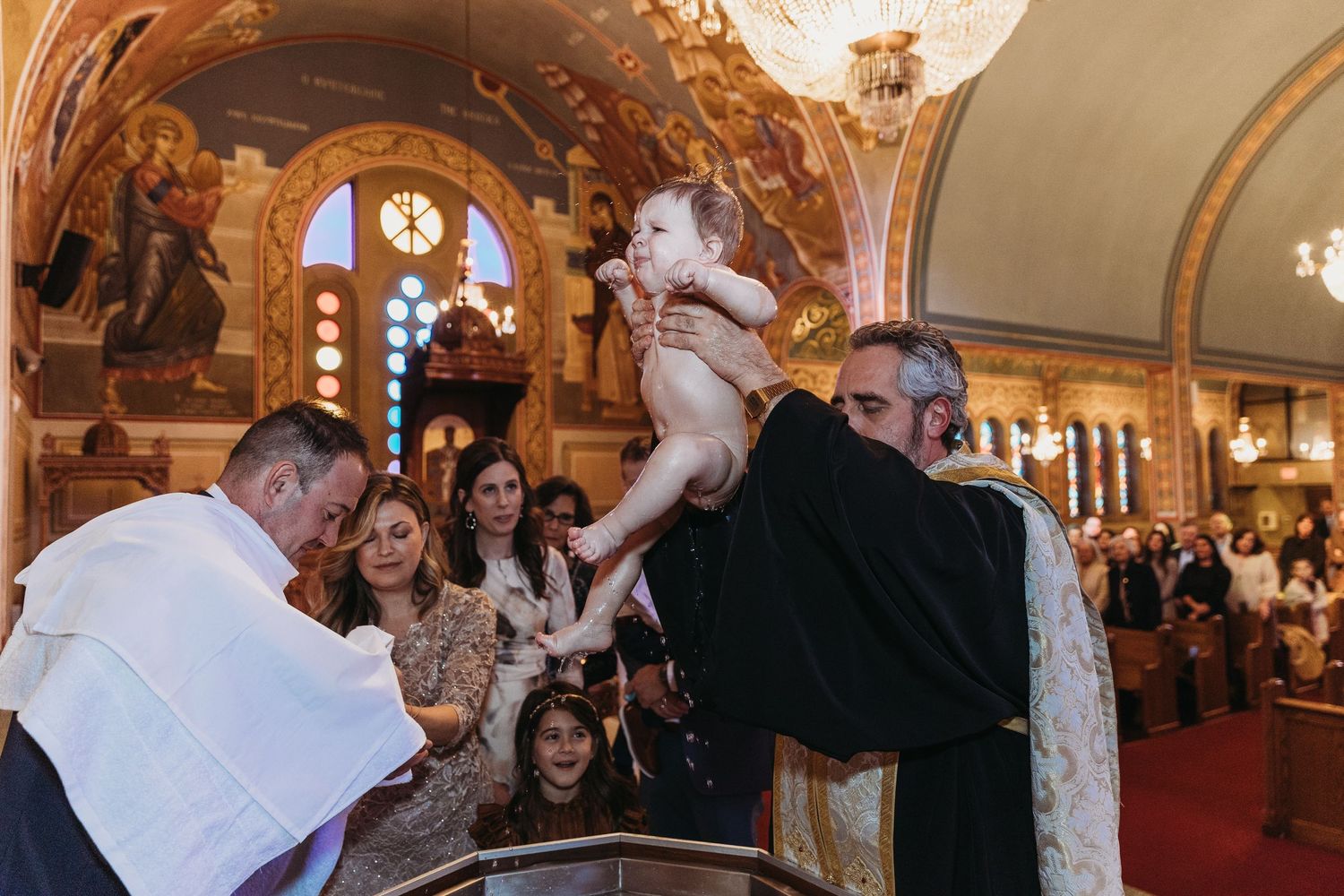 A priest holds a baby during a baptism ceremony inside an ornate Orthodox church with golden decor.