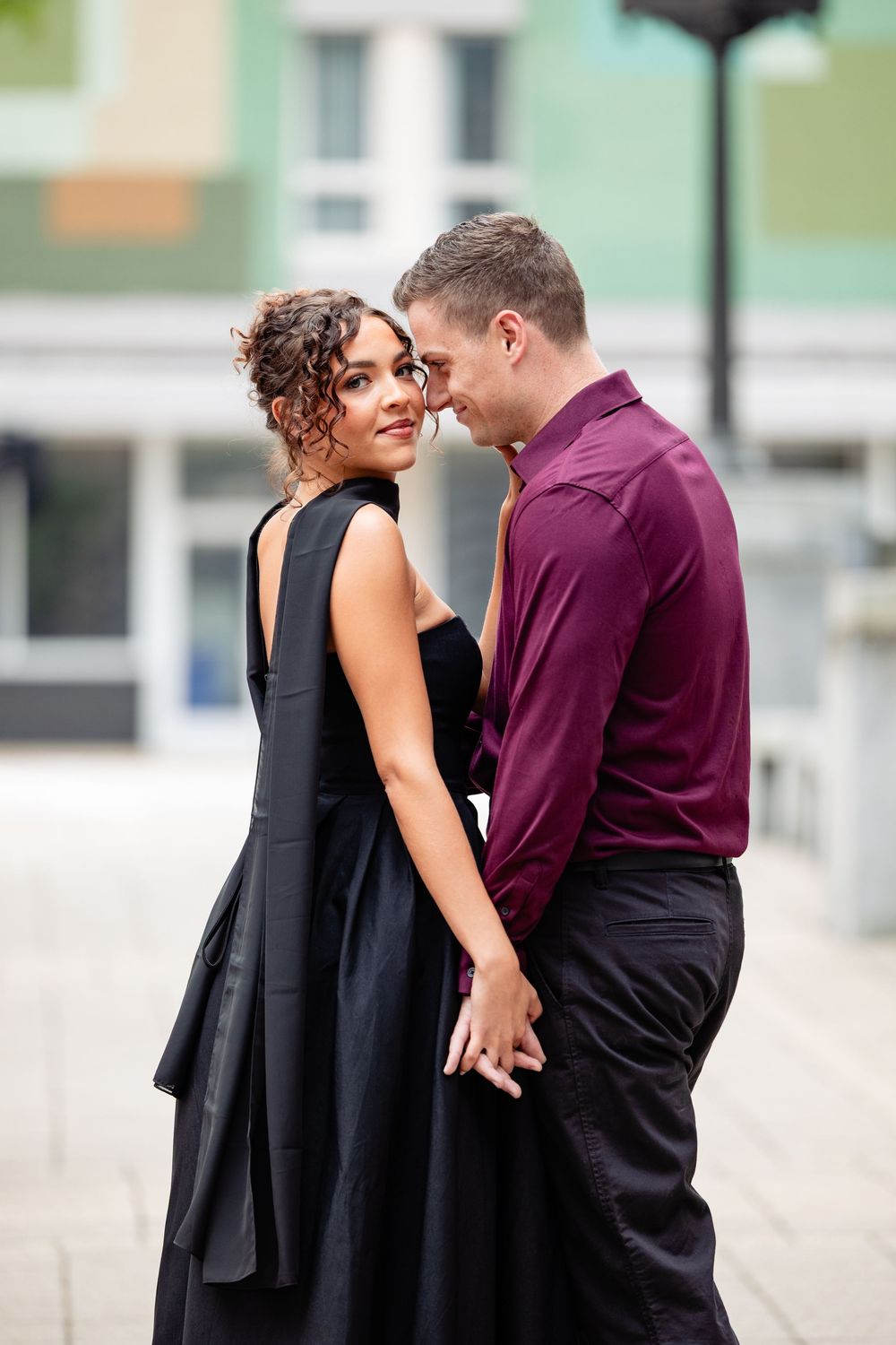Elegant couple in formal attire holding hands outdoors, woman in black dress smiling, man in burgundy shirt.