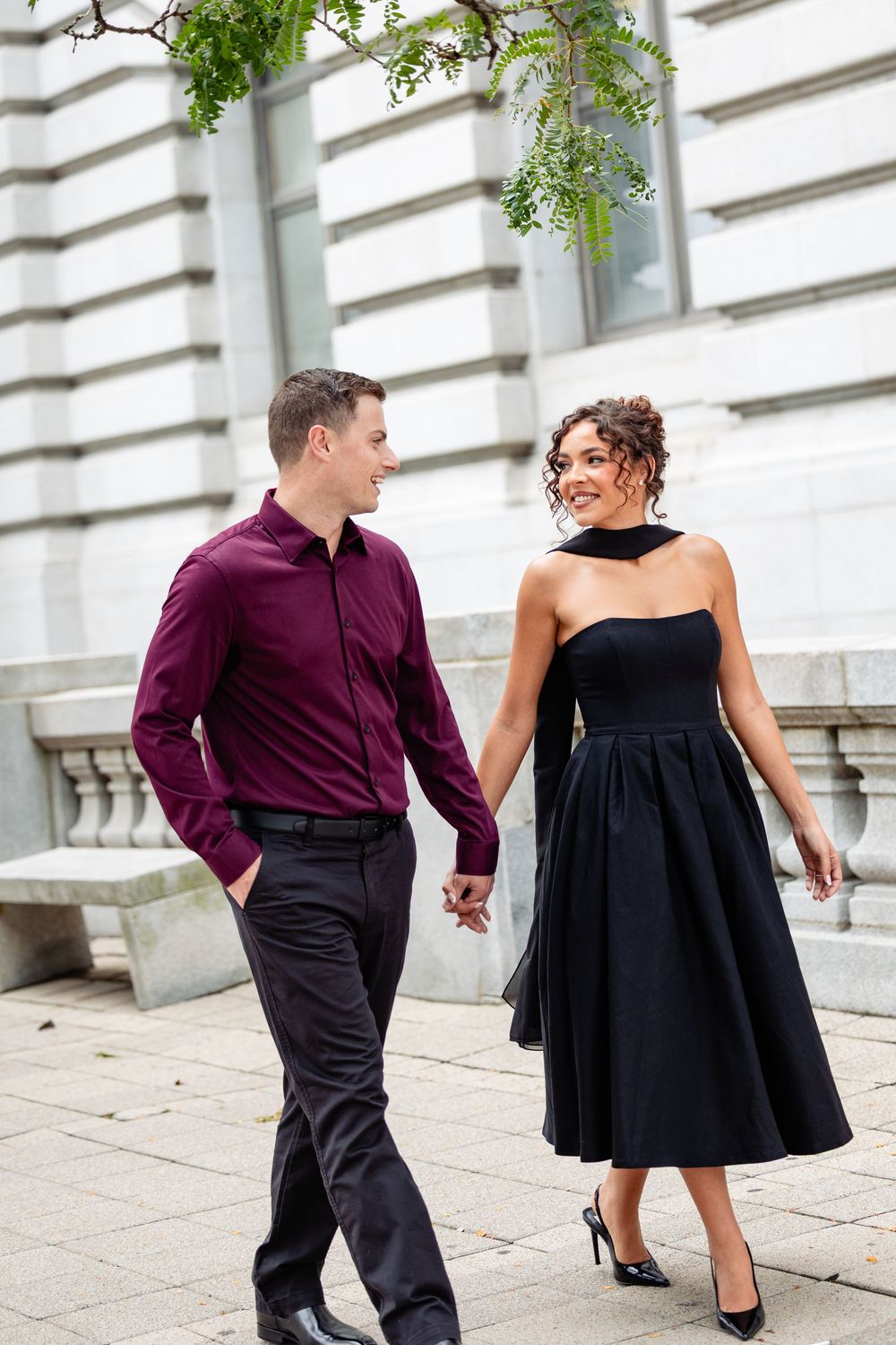 Elegant couple holding hands, man in burgundy shirt, woman in black strapless dress, walking outside a classic building.