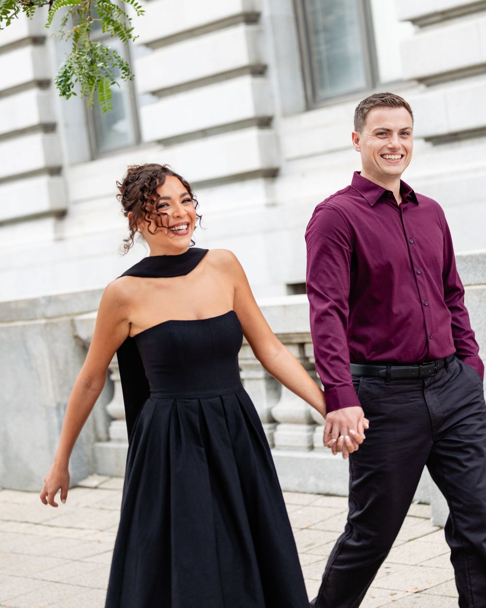 A smiling couple holding hands, woman in black dress and man in burgundy shirt, walking outdoors near a stone building.
