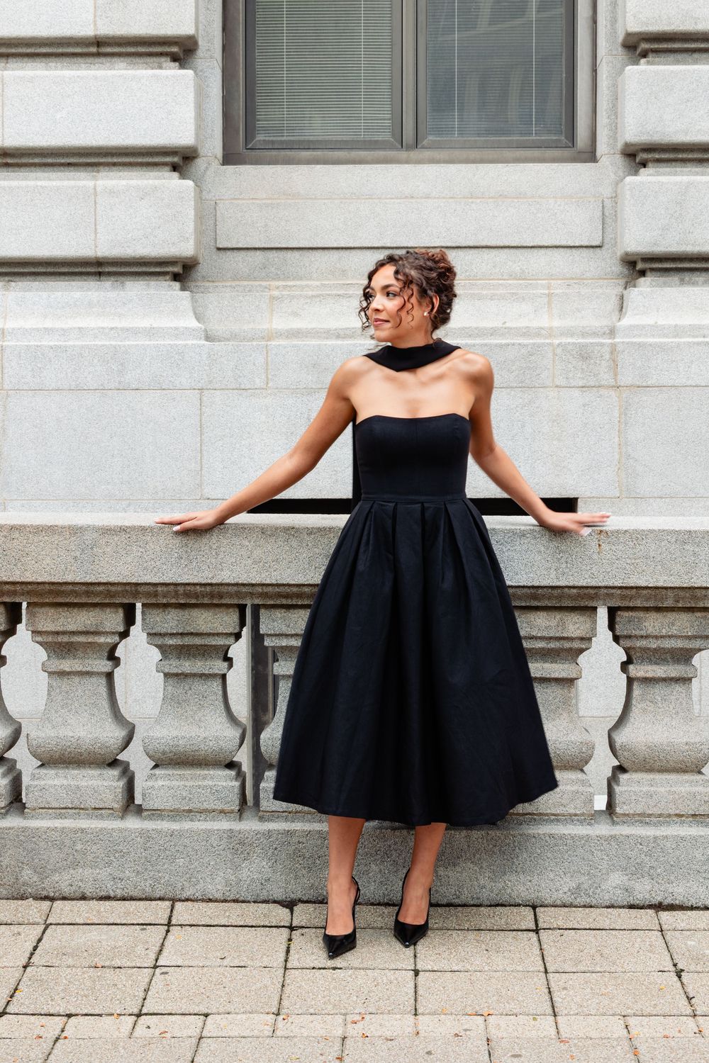Woman in black strapless midi dress with choker poses gracefully by stone balustrade.